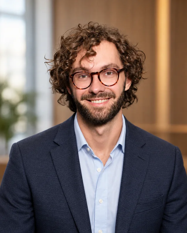 AI-generated man with glasses wearing a navy blazer and open-collar blue shirt against a softly blurred office background
